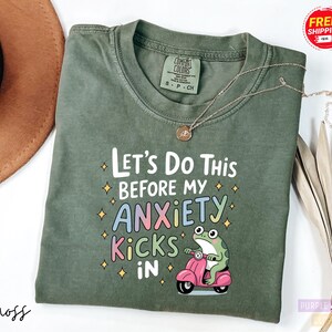 A green t-shirt with a colorful graphic and text printed on it, along with a small frog on a pink motorcycle.