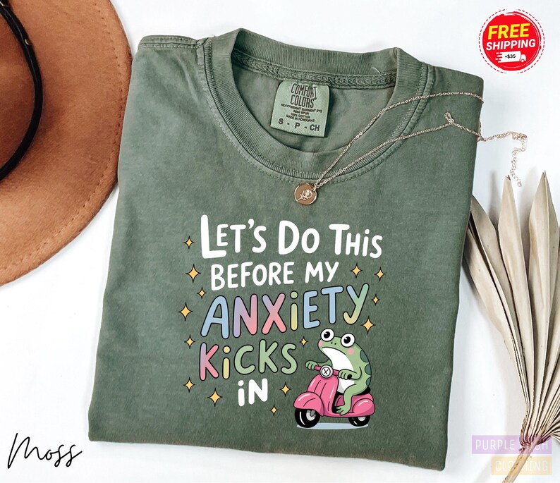 A green t-shirt with a colorful graphic and text printed on it, along with a small frog on a pink motorcycle.