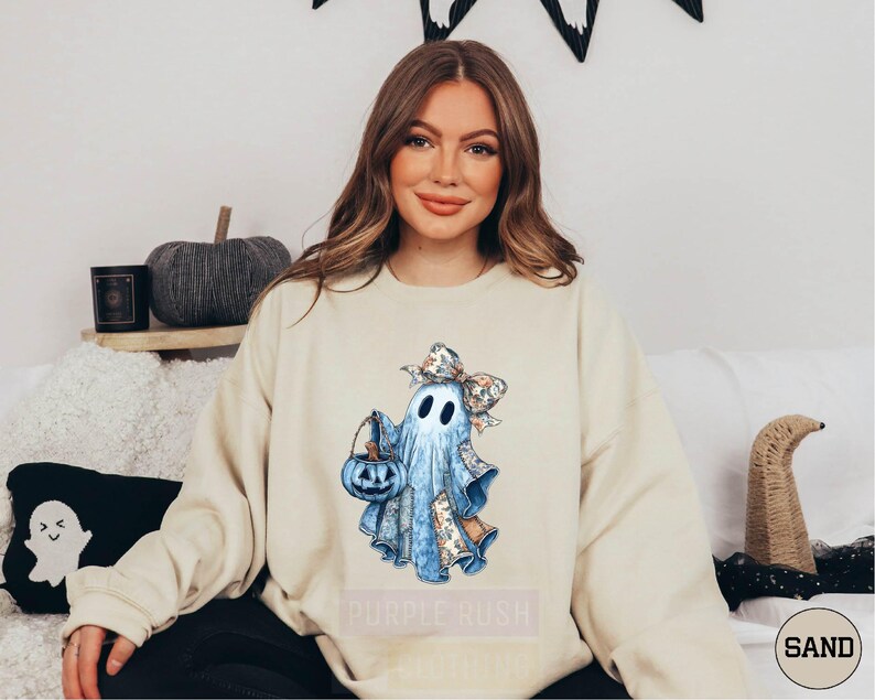 A woman wearing a white sweatshirt with a graphic of a ghost holding a pumpkin and a witch's hat, sitting on a couch with a pumpkin and a candle on a shelf behind her.