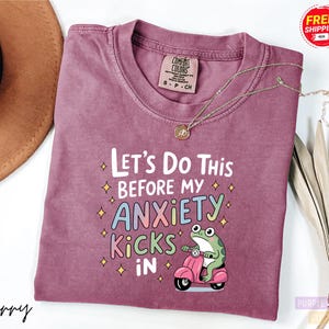 A purple t-shirt with a humorous text and a cartoon frog graphic, along with a necklace and a hat, are displayed on a white background.
