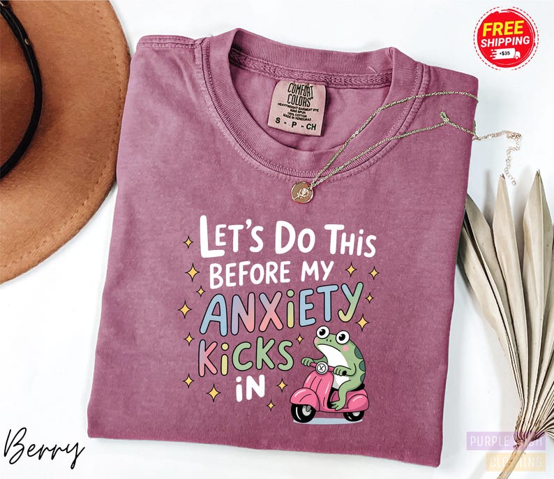 A purple t-shirt with a humorous text and a cartoon frog graphic, along with a necklace and a hat, are displayed on a white background.