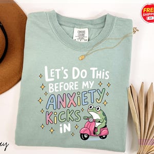 A light green t-shirt with a colorful graphic design and text, along with a brown hat and some decorative items, such as a necklace and a fan, are displayed on a white background.