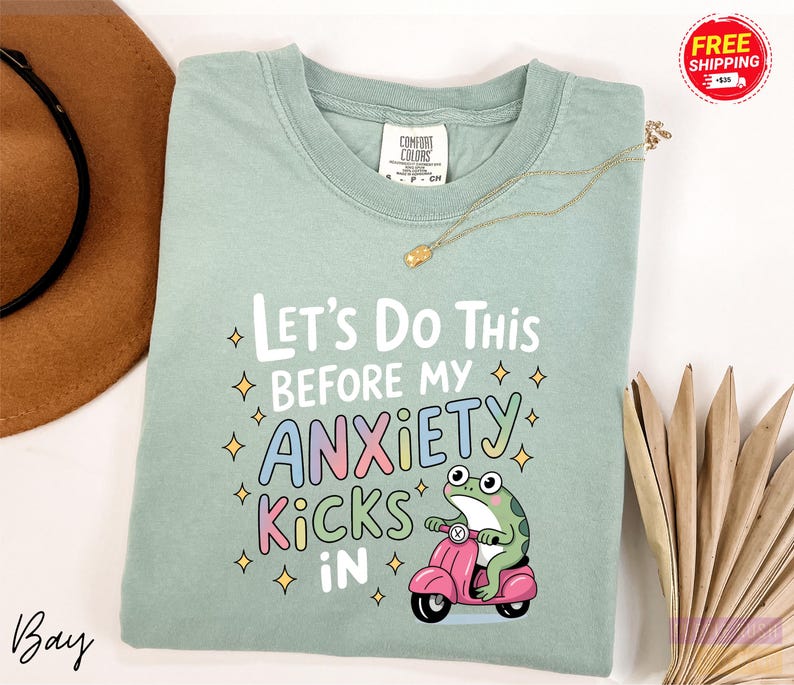 A light green t-shirt with a colorful graphic design and text, along with a brown hat and some decorative items, such as a necklace and a fan, are displayed on a white background.