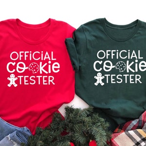May include: Two t-shirts, one red and one forest green, with the text "OFFICIAL COOKIE TESTER" in white. The design includes a cookie and gingerbread man graphic. The shirts are displayed with denim and holiday greenery.
