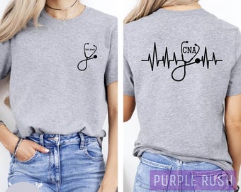 Op de afbeelding: Grijs T-shirt met een stethoscoop-afbeelding aan de voorkant en een hartslag-afbeelding met de tekst "CNA" aan de achterkant. Het shirt wordt getoond op een model dat een blauwe jeans draagt.