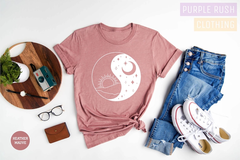 May include: A heather mauve t-shirt with a white sun and moon yin yang design. The shirt is laid flat on a white surface with a wooden cutting board, a camera, glasses, a wallet, a pair of jeans, and white sneakers. The text "Purple Rush Clothing" is visible in the top right corner of the image.