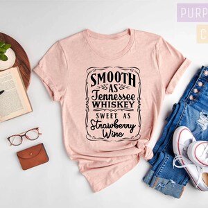 Op de afbeelding: Een heideperzikkleurig t-shirt met een zwarte afbeelding waarop staat "Smooth as Tennessee Whiskey Sweet as Strawberry Wine". Het shirt ligt op een wit oppervlak met een spijkerbroek, een boek, een bril en een kleine bruine portemonnee.