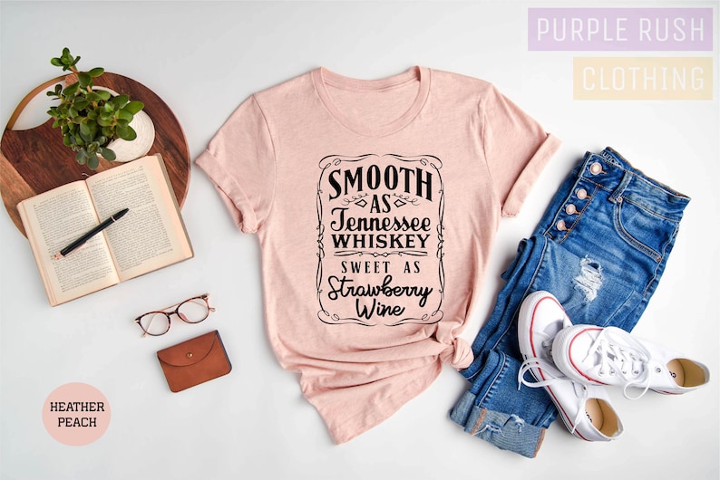 Op de afbeelding: Een heideperzikkleurig t-shirt met een zwarte afbeelding waarop staat "Smooth as Tennessee Whiskey Sweet as Strawberry Wine". Het shirt ligt op een wit oppervlak met een spijkerbroek, een boek, een bril en een kleine bruine portemonnee.