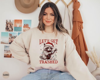 May include: Sand-colored sweatshirt with a raccoon wearing a Santa hat and Christmas lights. The text "LET'S GET TRASHED" is printed in red. The sweatshirt is a casual, festive garment.