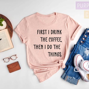 Puede incluir: Una camiseta rosa con el texto "FIRST I DRINK THE COFFEE, THEN I DO THE THINGS." en ella. La camiseta est&aacute; colocada sobre una superficie blanca con un par de vaqueros, un libro, gafas y una cartera.