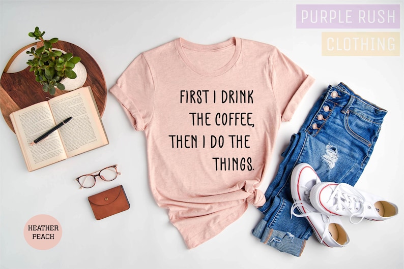 Puede incluir: Una camiseta rosa con el texto "FIRST I DRINK THE COFFEE, THEN I DO THE THINGS." en ella. La camiseta est&aacute; colocada sobre una superficie blanca con un par de vaqueros, un libro, gafas y una cartera.