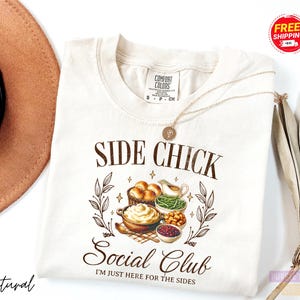 May include: Cream-colored t-shirt with the text "SIDE CHICK Social Club" and the phrase "I'M JUST HERE FOR THE SIDES." The graphic features illustrations of Thanksgiving side dishes. A gold necklace and a brown hat are also visible.