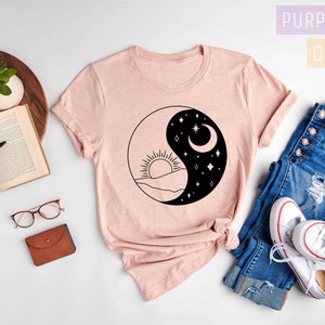 May include: A pink t-shirt with a black and white yin yang design featuring a sun and moon. The design also includes mountains and stars. The t-shirt is laid out on a white surface with a book, glasses, a wallet, a plant, and a pair of jeans and white sneakers.