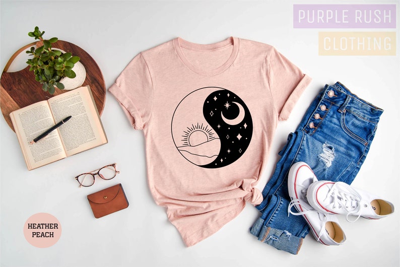 May include: A pink t-shirt with a black and white yin yang design featuring a sun and moon. The design also includes mountains and stars. The t-shirt is laid out on a white surface with a book, glasses, a wallet, a plant, and a pair of jeans and white sneakers.