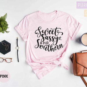 K&ouml;nnte beinhalten: Rosa T-Shirt mit dem Text "Sweet and Sassy Southern" in schwarzer Schrift. Das Shirt liegt flach auf einer wei&szlig;en Oberfl&auml;che mit einem Farn, einer Brille, einer Uhr, einem kleinen schwarzen Tablett, einer braunen Handtasche und einer Tasse Kaffee.