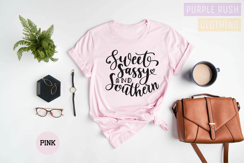 K&ouml;nnte beinhalten: Rosa T-Shirt mit dem Text "Sweet and Sassy Southern" in schwarzer Schrift. Das Shirt liegt flach auf einer wei&szlig;en Oberfl&auml;che mit einem Farn, einer Brille, einer Uhr, einem kleinen schwarzen Tablett, einer braunen Handtasche und einer Tasse Kaffee.