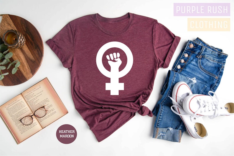 May include: A maroon t-shirt with a white feminist fist symbol. The shirt is laid flat on a white surface with a book, a wooden tray, and a pair of jeans and white sneakers.