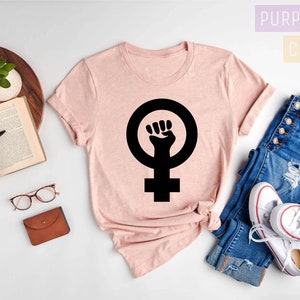 May include: A pink t-shirt with a black feminist fist symbol on a white background. The text "Purple Rush Clothing" is in the top right corner of the image. The text "Heather Peach" is in the bottom left corner of the image.