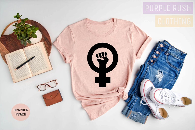 May include: A pink t-shirt with a black feminist fist symbol on a white background. The text "Purple Rush Clothing" is in the top right corner of the image. The text "Heather Peach" is in the bottom left corner of the image.