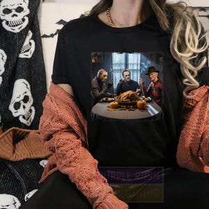 May include: Black t-shirt featuring a graphic of horror movie characters at a Thanksgiving dinner table. The shirt is worn by a person with a rust-colored cardigan. A black and white skull-patterned blanket is visible in the background.