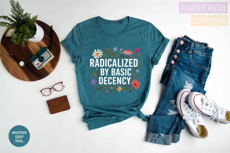 a casual outfit consisting of a teal t-shirt with a floral design, a pair of blue jeans, white sneakers, and a pair of glasses. The outfit is laid out on a white background, with a potted plant and a camera visible in the background.
