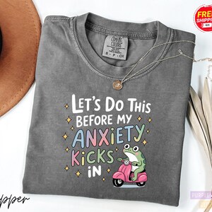A gray t-shirt with a colorful graphic design and text, along with a brown hat and some dried leaves, on a white background.