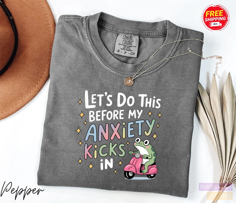 A gray t-shirt with a colorful graphic design and text, along with a brown hat and some dried leaves, on a white background.