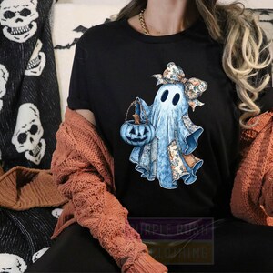 A person wearing a black t-shirt with a graphic of a ghost holding a pumpkin and a bow, sitting on a couch with a skull and spider web patterned blanket behind them.