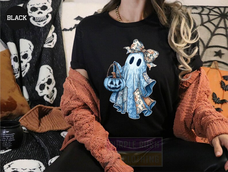 A person wearing a black t-shirt with a graphic of a ghost holding a pumpkin and a bow, sitting on a couch with a skull and spider web patterned blanket behind them.