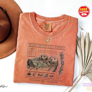 May include: A coral-colored t-shirt featuring a detailed graphic of sea otters and text about their habitat and diet. The shirt includes the text "Sea Otter Enhydra Lutris" and "Monterey Bay Aquarium". A gold necklace and a brown hat are also visible.