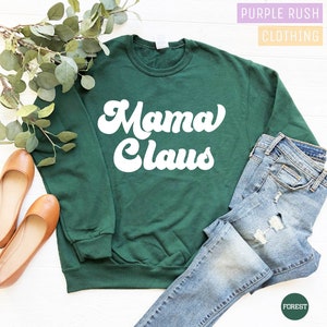 May include: A forest green sweatshirt with the words "Mama Claus" in white, retro-style lettering. The sweatshirt is paired with light-wash denim jeans and brown flats. The text "Purple Rush Clothing" is in the top right corner.