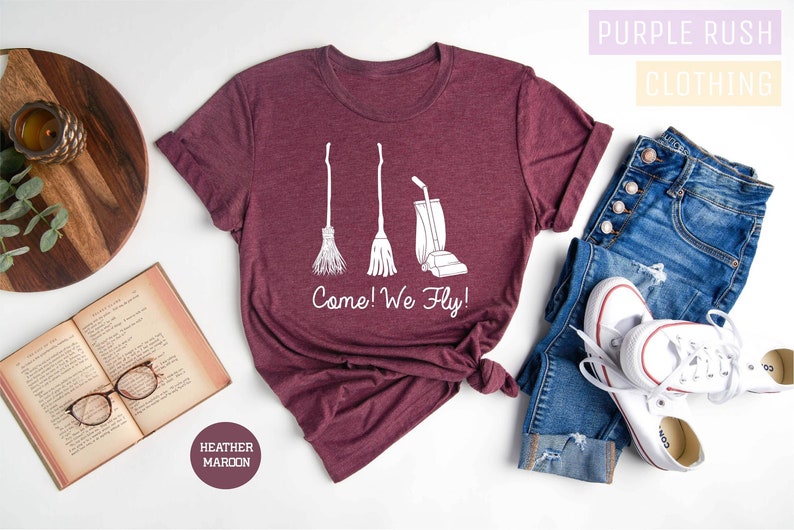 May include: Heather maroon t-shirt with a white graphic of a broom, a mop, and a vacuum cleaner, with the text "Come! We Fly!". The shirt is styled with blue jeans and white sneakers.