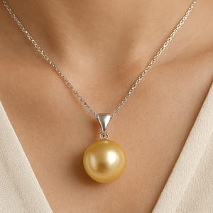 Puede incluir: Un collar de plata con un gran colgante de perla dorada. La perla es redonda y tiene un brillo lustroso. La cadena es delicada y cuelga de una fianza de plata.
