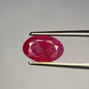 May include: A faceted, oval ruby gemstone, held by silver tweezers. The ruby is a deep red colour with internal reflections. The gemstone is displayed against a neutral grey background.