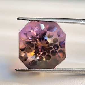 Könnte beinhalten: Ein facettierter Edelstein, der von einer Pinzette gehalten wird. Der Stein hat eine achteckige Form mit einem komplexen Innenmuster aus Kreisen und Facetten. Die Farben gehen von Lila zu Gold über.