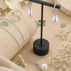 May include: A jewelry set consisting of a silver chain necklace with a single teardrop-shaped white pearl pendant and a pair of matching silver hoop earrings, each featuring a dangling teardrop-shaped white pearl, displayed on a black stand.