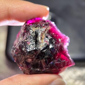 Op de afbeelding: Een ruwe, ongeslepen edelsteen met een diepe magenta tint, gecontrasteerd door donkere, bijna zwarte gebieden. Het oppervlak van de steen is getextureerd en onregelmatig, met een kristallijn uiterlijk. De edelsteen wordt tussen vingers gehouden.