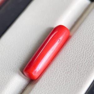 May include: A vibrant red, cylindrical object with rounded ends, possibly a decorative element or component. It rests on a textured, off-white surface, with a dark border visible. The object's smooth surface reflects light, highlighting its color.