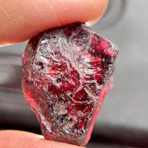 Puede incluir: Una gema en bruto, de color rojo intenso, con una estructura cristalina. La piedra tiene un color base oscuro, casi negro, con reflejos rojos brillantes. La superficie es irregular y texturizada, con facetas visibles y un aspecto natural sin pulir.