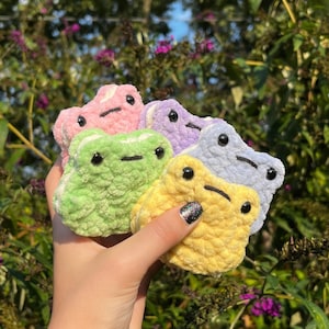 May include: Four crocheted frog plushies in different pastel colors: pink, purple, green, and yellow. The plushies are all the same size and have black eyes and a black mouth.