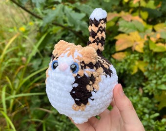 May include: A crocheted stuffed cat toy with a white, brown, and orange calico pattern. The cat has blue eyes and a small pink nose.
