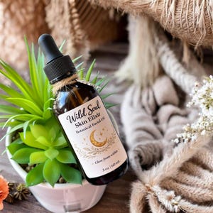 Puede incluir: Una botella de vidrio marrón de Wild Soul Skin Elixir Botanical Facial Oil con un cuentagotas negro y una etiqueta con una luna creciente y estrellas. La botella está envuelta con hilo y se encuentra frente a una pequeña planta en maceta. La botella contiene 60 ml.