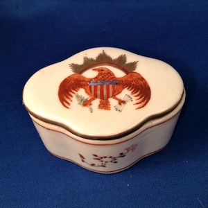 May include: A small, white porcelain trinket box with a lid. The lid is decorated with a hand-painted image of an eagle with outstretched wings, holding a bundle of arrows in its talons. The eagle is perched on a shield with red and white stripes, and a blue band across the top. The box is trimmed with gold paint.