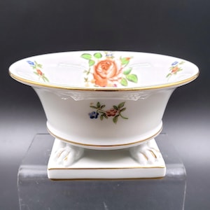 Peut inclure: Un pot à fleurs en porcelaine blanche avec un bord doré et un motif floral. Le pot a trois pieds en forme de pattes de lion et repose sur une base carrée.