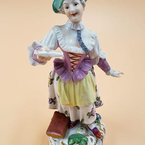 Könnte beinhalten: Porzellanfigur einer Frau in traditioneller Kleidung, die einen Teller hält. Die Figur trägt einen grünen Hut, eine weiße Bluse, ein lilafarbenes Korsett und einen Rock mit Blumenmuster. Die Figur steht auf einem dekorativen Sockel mit grünem Laub.