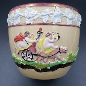 May include: A beige ceramic planter featuring a raised relief design of two cherubs, one riding in a small cart and the other riding a feline creature. The rim is decorated with a white grape vine pattern and a purple band, with green foliage at the base.