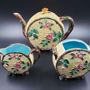 May include: A three-piece ceramic tea set with a basket weave design. The set includes a teapot, a creamer, and a sugar bowl. Each piece is decorated with pink flowers and green leaves. The interior of the creamer and sugar bowl is turquoise.