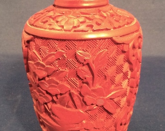 May include: A small, red, carved vase with a floral and bird design. The vase has a gold rim and is made of lacquered wood.
