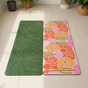 May include: Two yoga mats with colorful designs. One mat is green with a gold, flowing pattern. The other is pink with orange, yellow, and pink floral designs. The mats are on a light wood floor, with plants in the background.