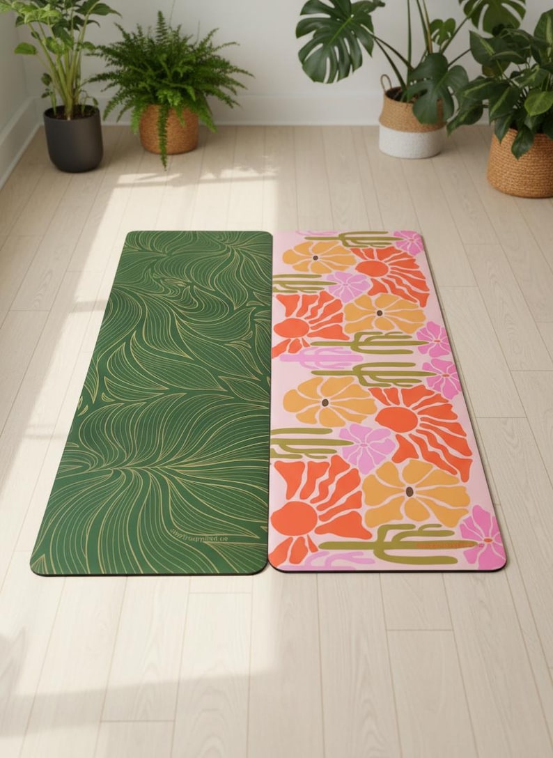 May include: Two yoga mats with colorful designs. One mat is green with a gold, flowing pattern. The other is pink with orange, yellow, and pink floral designs. The mats are on a light wood floor, with plants in the background.